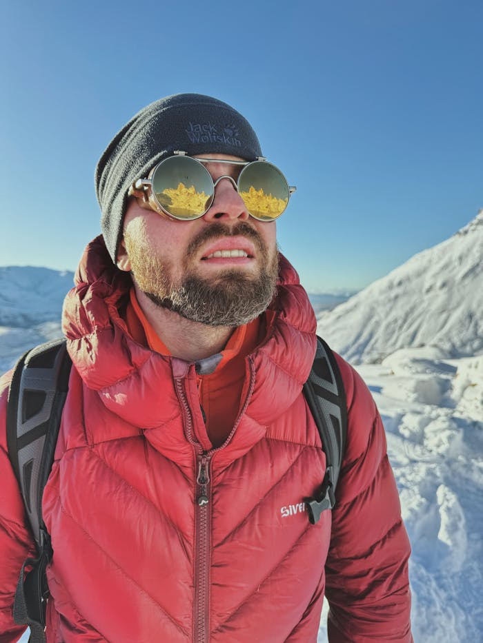 Home Man in a red jacket hiking in snowy mountains with sunglasses and backpack.