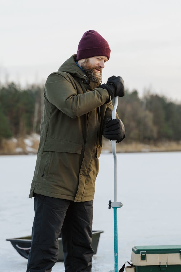 Bearded man wearing winter clothing, ice fishing on a frozen lake.